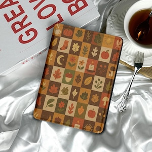 以下が含まれることがあります： 秋をテーマにしたデザインのタブレットケース。ケースは、葉、カボチャ、ドングリなどの秋に関連するイラストが描かれた、茶色、オレンジ、ベージュの色合いの正方形のパッチワークパターンが特徴です。ケースは白いサテンの表面にあります。