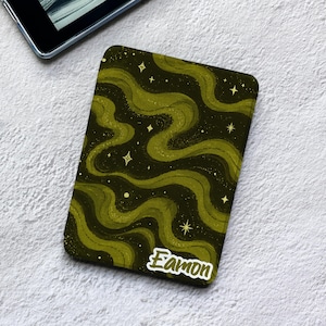 May include: A rectangular electronic reader cover featuring a dark olive green and black wavy pattern with scattered four-pointed stars and small dots. The name Eamon is printed in a white, stylized script font in the bottom right corner.