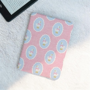 May include: Pink e-reader cover featuring a pattern of white geese in blue oval frames. Each goose has a pink bow. The cover is next to a black e-reader.