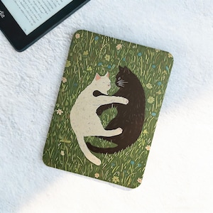 May include: A rectangular, green cat-themed decorative item featuring a white cat embracing a black cat, set against a backdrop of grass and flowers. The item is placed on a white surface, with a Kindle e-reader visible in the upper left corner.