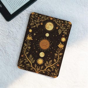 May include: A black e-reader cover with a celestial design featuring a sun, moons, stars, and floral elements in gold. The cover is rectangular and has rounded corners. An e-reader is partially visible in the upper left corner.