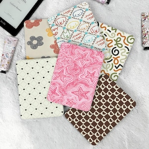 May include: A collection of colorful Kindle covers with various patterns, including flowers, hearts, stars, and puppies. The covers are in shades of pink, brown, cream, and white. A Kindle e-reader and a tube of lotion are also visible.
