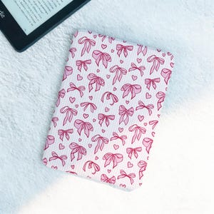 May include: A pink and white Kindle cover with a pattern of pink bows and hearts. The cover is rectangular and designed to protect an e-reader. The background is a soft, white, textured surface.