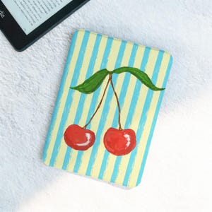 Op de afbeelding: Een rechthoekige hoes met een geschilderd kersendesign. De hoes heeft een lichtgele achtergrond met verticale blauwe strepen. Twee rode kersen met groene bladeren zijn op de voorkant geschilderd. Een e-reader is gedeeltelijk zichtbaar in de linkerbovenhoek.