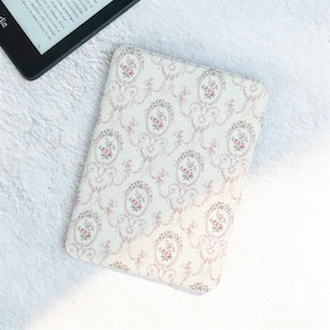 Può includere: Una custodia per Kindle con motivo floreale, con sfondo bianco e design floreale rosa. La custodia presenta un motivo ripetuto di cornici ovali con rose rosa e foglie verdi. Un e-reader è accanto.