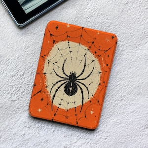 Könnte beinhalten: Eine rechteckige E-Reader-Hülle mit leuchtend orangefarbenem Hintergrund und einem cremefarbenen Kreis in der Mitte. Eine schwarze Spinne sitzt in der Mitte eines detaillierten schwarzen Spinnennetzes, das die gesamte Oberfläche bedeckt.