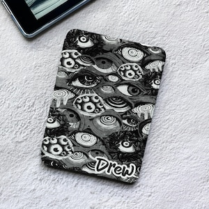 Può includere: Una custodia rettangolare per tablet con un motivo grafico in bianco e nero composto da numerosi occhi sovrapposti con vari design dell'iride. La parola Drew è stampata in un carattere bianco stilizzato in stile manoscritto sul fondo.