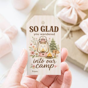 May include: A gift tag with the text "SO GLAD you wandered into our camp." The tag features a watercolour illustration of a lantern, flowers, a campfire, and a small wooden sign. The tag is held by a hand.