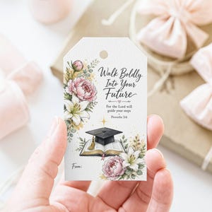 May include: A white gift tag with floral illustrations and the text "Walk Boldly Into Your Future." The tag features a graduation cap over an open book, with pink peonies and white lilies. The tag is held by a hand.