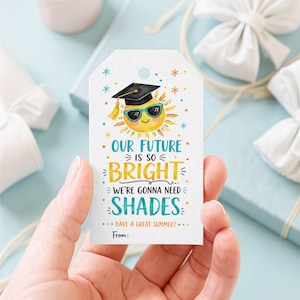 Peut inclure: Une étiquette cadeau blanche sur le thème de la remise de diplômes. L'étiquette représente un soleil souriant portant des lunettes de soleil et un chapeau de remise de diplômes. Le texte sur l'étiquette se lit : « OUR FUTURE IS SO BRIGHT, WE'RE GONNA NEED SHADES ». L'étiquette est tenue par une main.