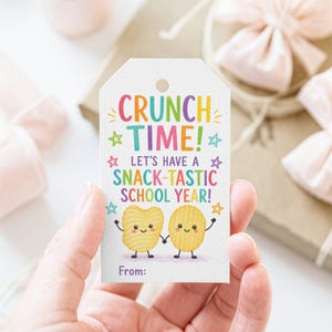 May include: A white rectangular gift tag with a hole at the top. The text reads CRUNCH TIME! LET'S HAVE A SNACK-TASTIC SCHOOL YEAR! From: in colorful letters. Two cartoon potato chips with faces and arms are holding hands, surrounded by stars.