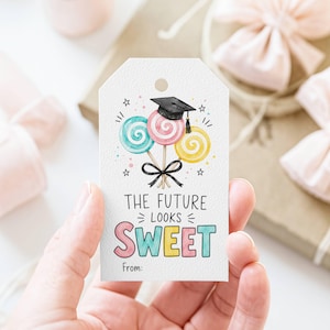 Peut inclure: Une étiquette cadeau blanche avec une illustration à l'aquarelle de trois sucettes, un chapeau de remise de diplôme et le texte "THE FUTURE LOOKS SWEET". Les sucettes sont bleues, roses et jaunes. Un nœud noir est noué sous les sucettes.