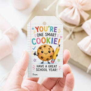 May include: A rectangular paper gift tag with a white background featuring blue horizontal lines. The text reads YOU'RE ONE SMART COOKIE! HAVE A GREAT SCHOOL YEAR! and From:. A cartoon chocolate chip cookie holds a yellow pencil with a pink eraser.