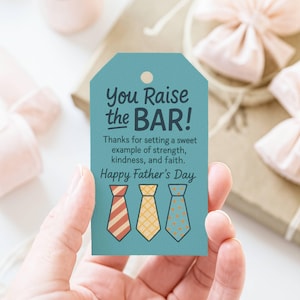 Könnte beinhalten: Ein türkisfarbenes Geschenk-Etikett mit dem Text "You Raise the BAR!" und "Happy Father's Day." Das Etikett zeigt drei illustrierte Krawatten in Rot, Gelb und Blau. Das Etikett wird in einer Hand gehalten.