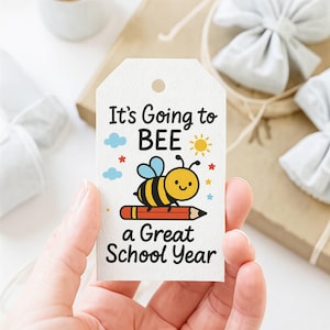 May include: A white rectangular gift tag with a hole at the top, featuring a cartoon bee sitting on an orange pencil. The text reads It's Going to BEE a Great School Year, accompanied by small blue clouds, a yellow sun, and red and blue stars.
