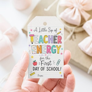 May include: A rectangular white gift tag with a hole at the top, featuring a lined paper background and colorful text that reads A Little Sip of TEACHER ENERGY for the First DAY OF SCHOOL! with illustrations of a pencil, apple, and paperclip.