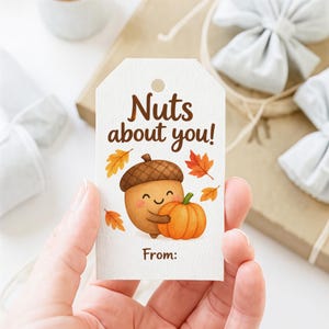 Op de afbeelding: Een wit rechthoekig cadeaulabel met een gat aan de bovenkant, voorzien van de bruine tekst "Nuts about you!" boven een cartoonillustratie van een lachende eikel die een oranje pompoen vasthoudt, omringd door kleine gele en oranje herfstbladeren.