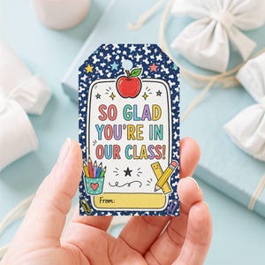 Può includere: Un'etichetta regalo rettangolare con sfondo blu scuro maculato, stelle colorate e illustrazioni a tema scolastico. Il testo centrale recita "SO GLAD YOU'RE IN OUR CLASS!" con una mela rossa, un portapenne e un righello. In basso c'è un campo giallo "From:" per il mittente.