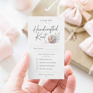 May include: A cream-colored card with the words "Lovingly Made Handcrafted Knit" in elegant script. A watercolor illustration of a yarn ball and foliage accents the design. The card includes fiber content and care instructions.