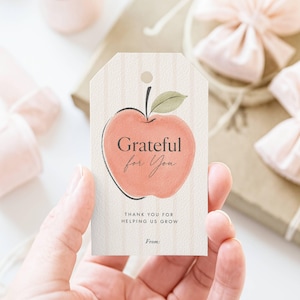Può includere: Un'etichetta regalo rettangolare in carta con un motivo a righe verticali beige chiaro. Al centro presenta un'illustrazione in stile acquerello di una mela color pesca con una foglia verde e il testo Grateful for You, THANK YOU FOR HELPING US GROW, From:.