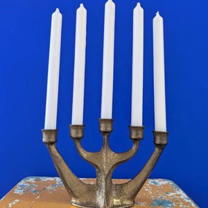Può includere: Un candelabro color ottone con cinque candele bianche. Il candelabro presenta un design stilizzato unico con bracci curvi. Lo sfondo è blu vibrante e il candelabro poggia su una superficie verniciata di giallo usurato.