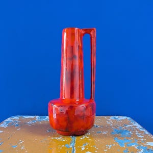 Op de afbeelding: Een hoge, rode keramische vaas met een handvat. De vaas heeft een cilindrische vorm en een afgeronde basis. Het rode glazuur heeft donkere, gevlekte accenten. De vaas staat op een verweerd geel en blauw oppervlak tegen een blauwe achtergrond.