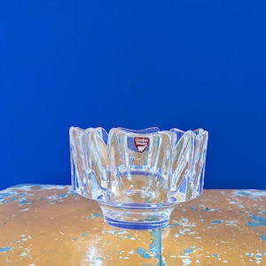 Op de afbeelding: Een heldere, facetgeslepen glazen kom met een brede, geschulpte rand. De kom heeft een cirkelvormige basis en een klein, rechthoekig label met de tekst "Orrefors Sweden". De kom wordt getoond op een verweerd, oranje en blauw oppervlak tegen een blauwe achtergrond.