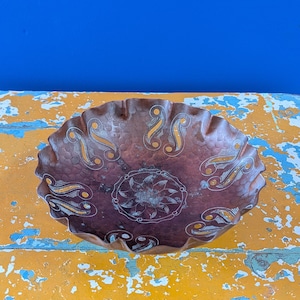 Op de afbeelding: Een decoratieve, gehamerde koperen kom met een gegolfde rand. De kom heeft een ingewikkeld ontwerp van bloemen- en paisley-patronen in goud en wit. De kom staat op een verweerd gele ondergrond en een blauwe achtergrond.