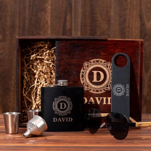 May include: A personalized gift set featuring a black flask, sunglasses, bottle opener, shot glasses, and a funnel. The flask and bottle opener are engraved with a monogram and the name "DAVID". The items are presented in a wooden box.