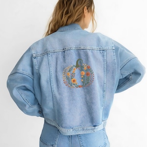 May include: Light blue denim jacket with a pumpkin design embroidered on the back. The pumpkin is composed of orange, yellow, and green floral elements, creating a fall-themed aesthetic. The jacket has a classic denim style with a button-up front.