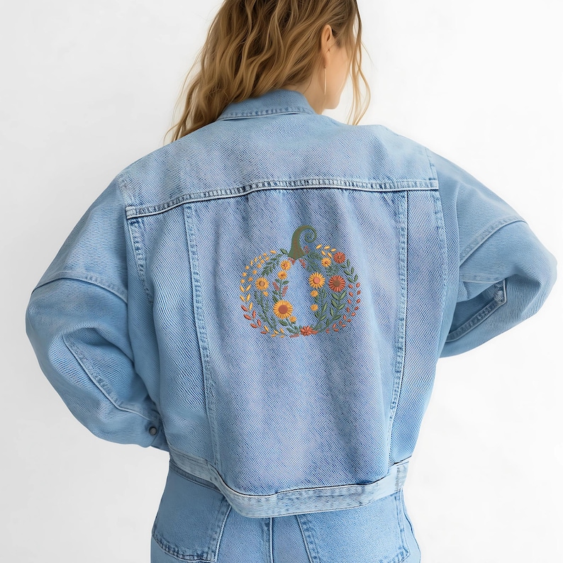 May include: Light blue denim jacket with a pumpkin design embroidered on the back. The pumpkin is composed of orange, yellow, and green floral elements, creating a fall-themed aesthetic. The jacket has a classic denim style with a button-up front.