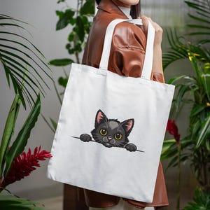 May include: White tote bag featuring a black cat graphic. The cat has yellow eyes, pink ears, and is peeking over a horizontal line. The bag has two white handles and is held by a person.