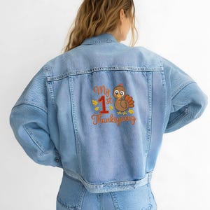 Op de afbeelding: Lichtblauwe spijkerjas met een geborduurd ontwerp op de rug. Het ontwerp toont een cartoon kalkoen, het nummer 1 en de woorden "My 1st Thanksgiving" in oranje en geel.