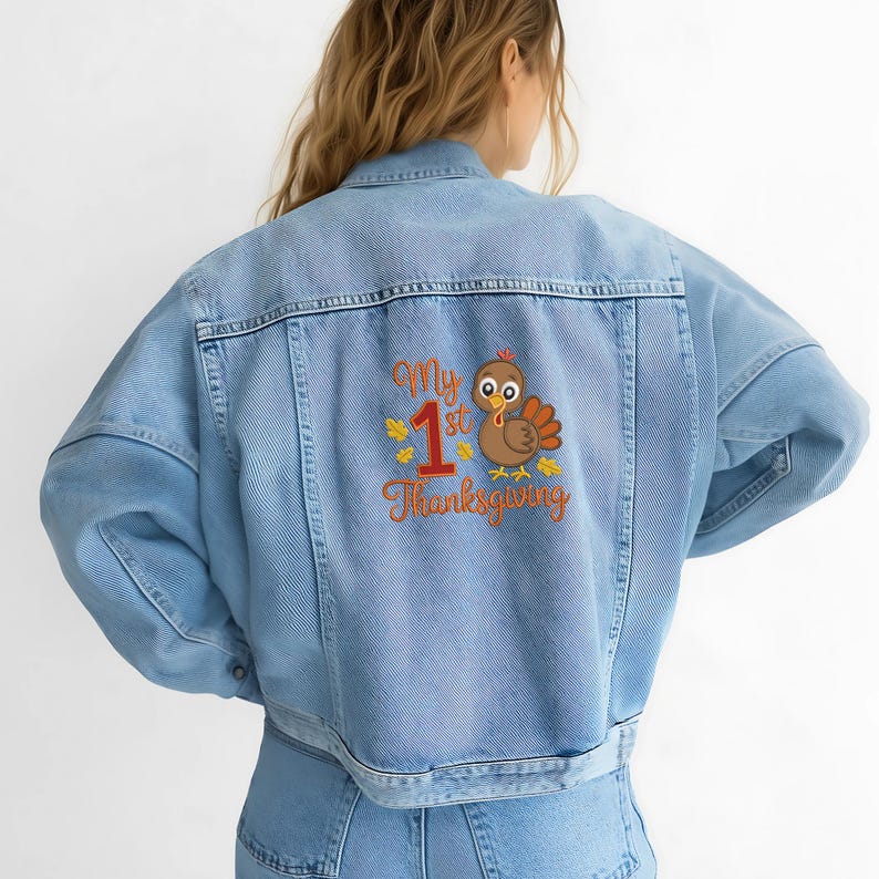 Op de afbeelding: Lichtblauwe spijkerjas met een geborduurd ontwerp op de rug. Het ontwerp toont een cartoon kalkoen, het nummer 1 en de woorden "My 1st Thanksgiving" in oranje en geel.