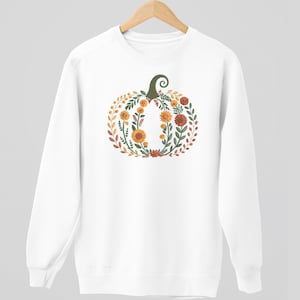 May include: White sweatshirt featuring a floral pumpkin design. The pumpkin is composed of orange, yellow, and red flowers, green leaves, and a green stem. The design is centered on the front of the sweatshirt, creating a fall-themed aesthetic.