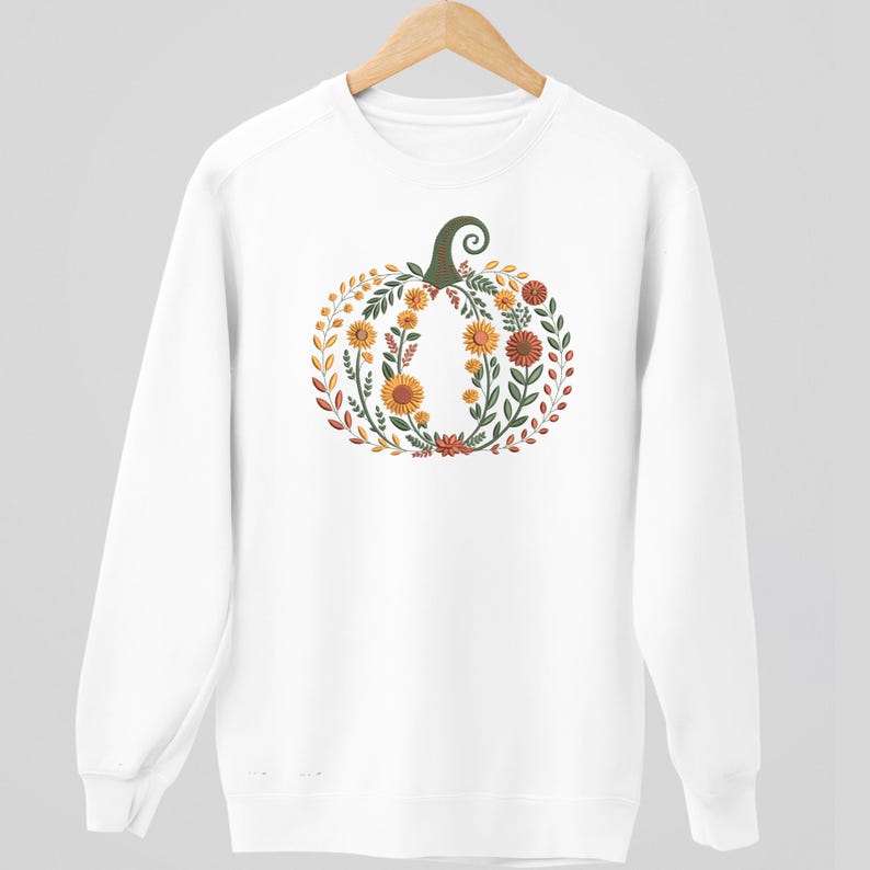 May include: White sweatshirt featuring a floral pumpkin design. The pumpkin is composed of orange, yellow, and red flowers, green leaves, and a green stem. The design is centered on the front of the sweatshirt, creating a fall-themed aesthetic.