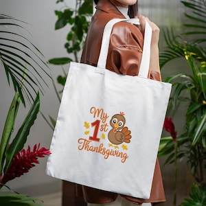 Op de afbeelding: Witte draagtas met de tekst "My 1st Thanksgiving" in oranje script en een cartoon kalkoen illustratie. De kalkoen is bruin met oranje en gele accenten. De tas heeft witte handvatten.