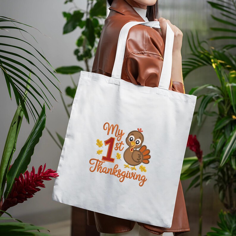 Op de afbeelding: Witte draagtas met de tekst "My 1st Thanksgiving" in oranje script en een cartoon kalkoen illustratie. De kalkoen is bruin met oranje en gele accenten. De tas heeft witte handvatten.