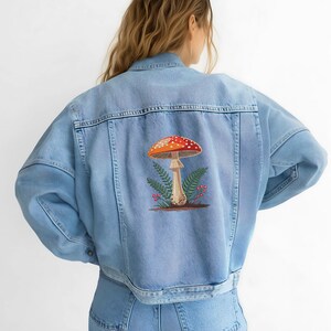 May include: Light blue denim jacket with a colorful mushroom design embroidered on the back. The mushroom has a red and white spotted cap, with green fern leaves and small red berries. The jacket has a classic button-up design.