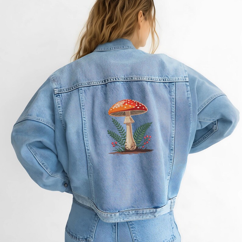 May include: Light blue denim jacket with a colorful mushroom design embroidered on the back. The mushroom has a red and white spotted cap, with green fern leaves and small red berries. The jacket has a classic button-up design.