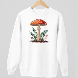 May include: White sweatshirt featuring a graphic of a red and white spotted mushroom with green ferns and red berries. The mushroom is centered on the front of the sweatshirt, creating a nature-inspired design.