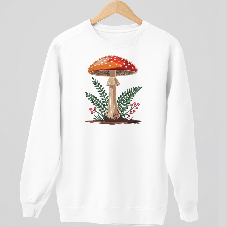 May include: White sweatshirt featuring a graphic of a red and white spotted mushroom with green ferns and red berries. The mushroom is centered on the front of the sweatshirt, creating a nature-inspired design.