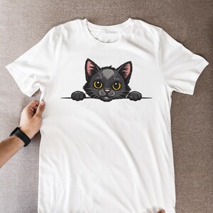 May include: White t-shirt featuring a cartoon black cat with yellow eyes and pink ears. The cat is peeking over a black line. The graphic is centered on the front of the shirt.