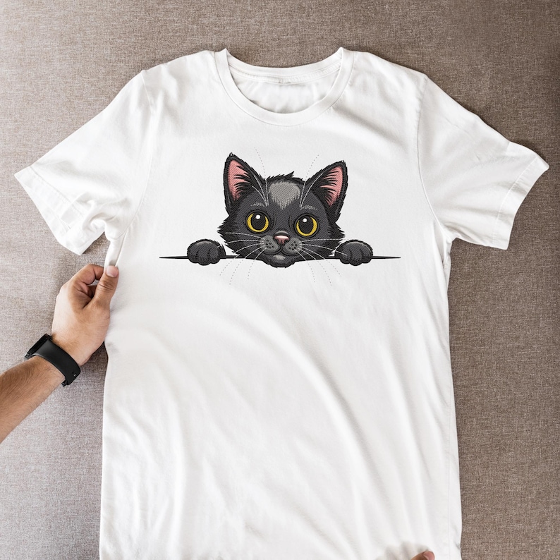 May include: White t-shirt featuring a cartoon black cat with yellow eyes and pink ears. The cat is peeking over a black line. The graphic is centered on the front of the shirt.