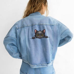 May include: Light blue denim jacket with a black cat graphic on the back. The cat has yellow eyes and is peeking over a line. The jacket has a classic design with pockets and button closures.