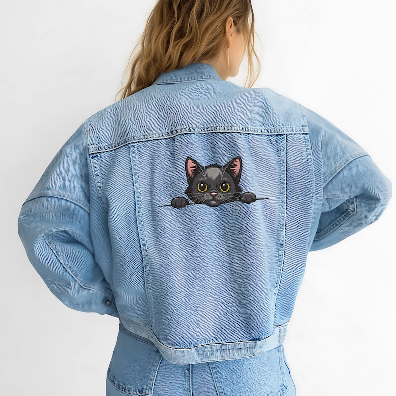 May include: Light blue denim jacket with a black cat graphic on the back. The cat has yellow eyes and is peeking over a line. The jacket has a classic design with pockets and button closures.