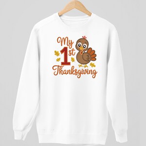 Op de afbeelding: Witte sweatshirt met een cartoon kalkoen afbeelding en de tekst "My 1st Thanksgiving" in oranje en rood. De kalkoen is bruin met oranje accenten en is omgeven door herfstbladeren.