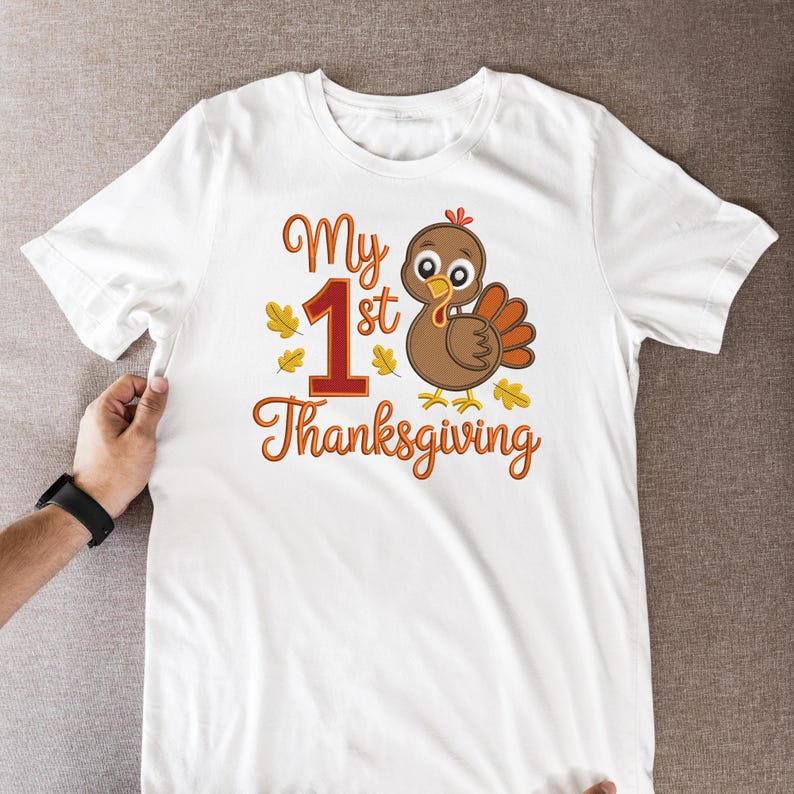 Op de afbeelding: Wit T-shirt met de tekst "My 1st Thanksgiving" in oranje script, een groot rood nummer 1 en een cartoon kalkoen. De kalkoen heeft bruine veren en een rode lellen. Gele bladeren zijn verspreid.