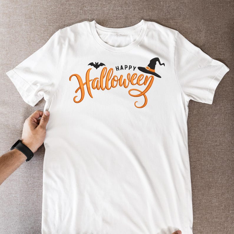 May include: White t-shirt with the words "Happy Halloween" in orange script, a black bat, and a witch's hat. The shirt is held up by a person's hand, showcasing the design. A black watch is visible on the wrist.