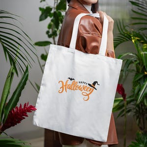 May include: A white tote bag with the words "Happy Halloween" in orange script, a black bat, and a witch's hat. The bag has white handles and is held by a person. The background features green plants and a brown jacket.